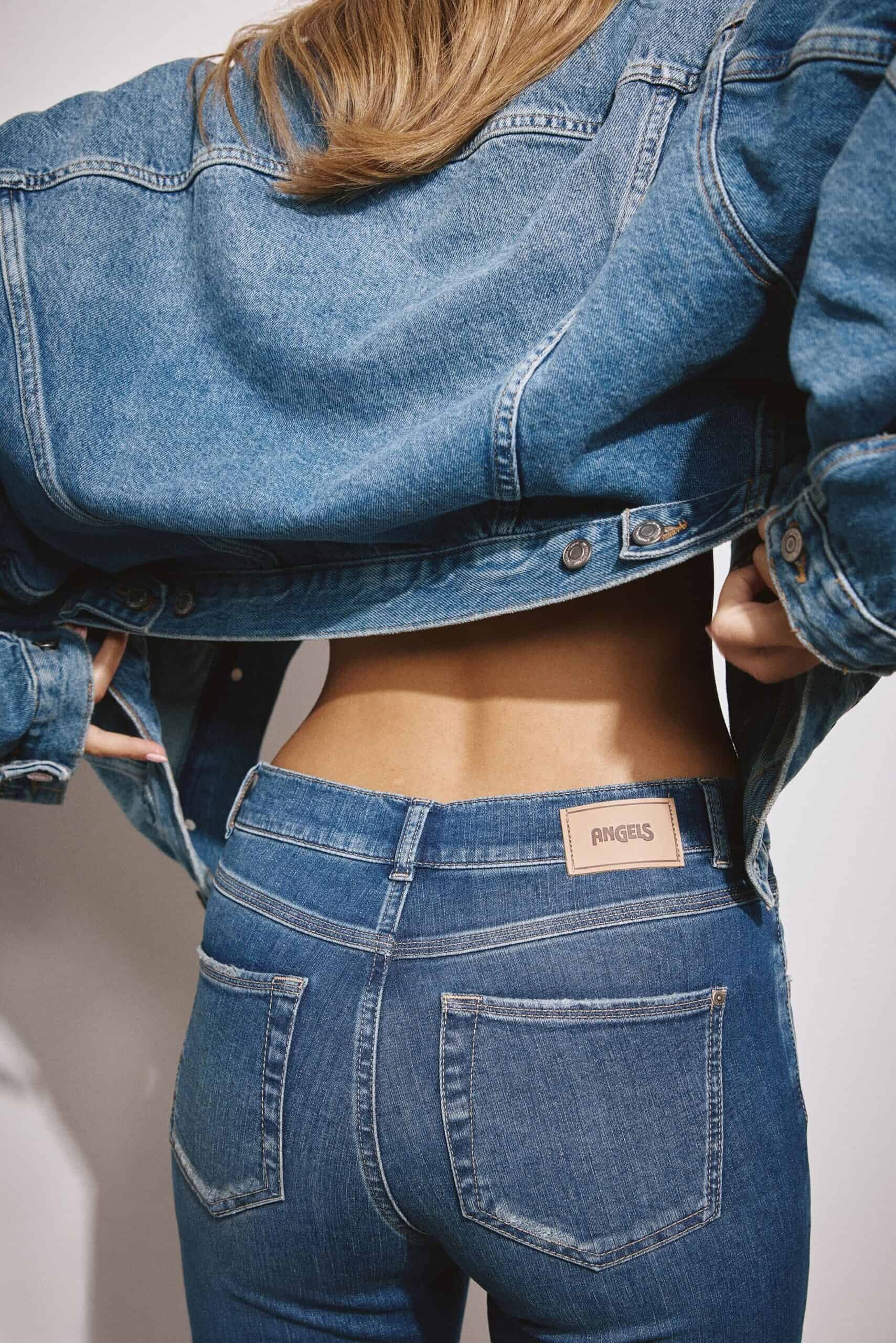 eine person, die eine blaue jeans und eine dazu passende jeansjacke trägt, von hinten gesehen, wobei sie die jacke anhebt, um ihren unteren rücken zu zeigen. die jeans hat ein etikett mit der aufschrift angels über der rechten tasche.