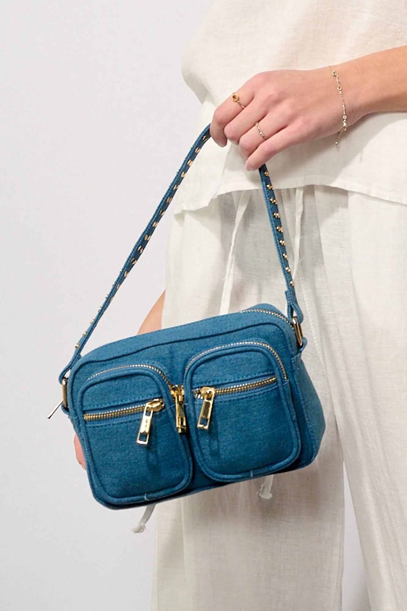 Eine weiß gekleidete Person hält eine blaugrüne Handtasche mit goldenen Reißverschlüssen und zwei Vordertaschen sowie einem kurzen, mit goldenen Nieten verzierten Henkel.