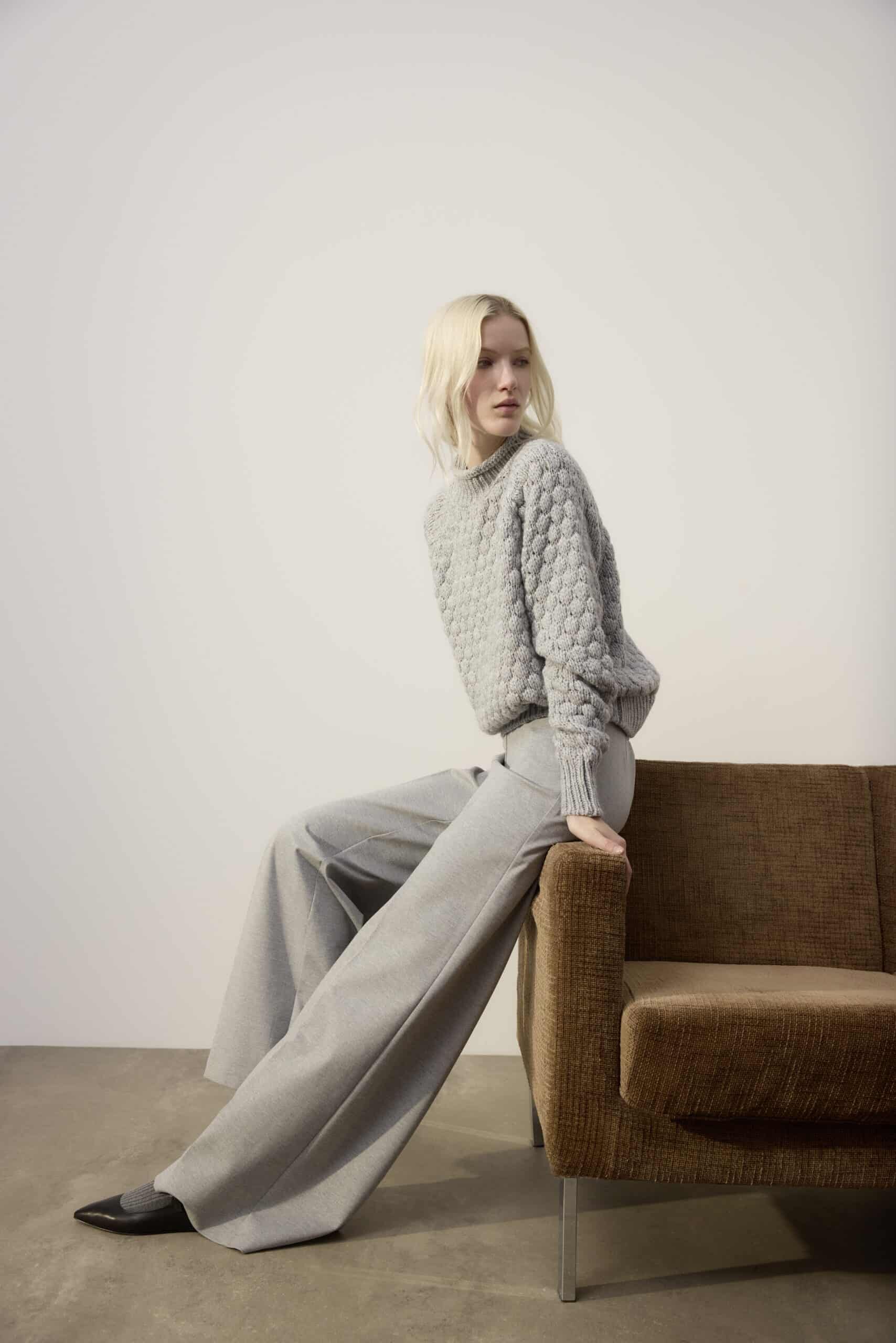 Eine Frau mit blondem Haar, die einen strukturierten grauen Pullover und eine graue Hose mit weitem Bein trägt, sitzt seitlich auf der Lehne eines braunen Sofas vor einer schlichten hellen Wand.