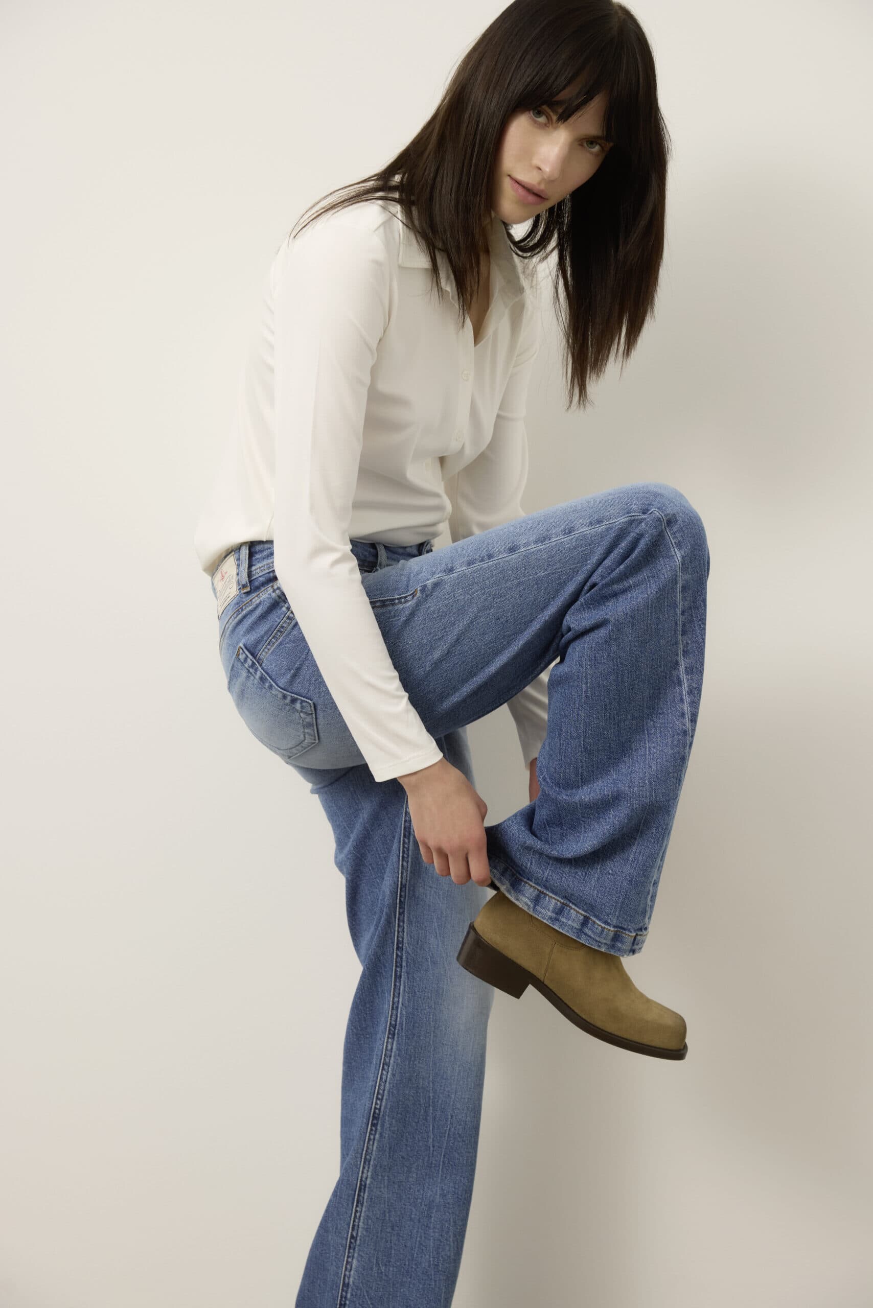 Eine Person mit glattem dunklem Haar, die ein weißes langärmeliges Oberteil und eine blaue Jeans trägt, beugt sich leicht nach vorne und richtet einen braunen Stiefel an ihrem erhobenen Fuß vor einem schlichten hellen Hintergrund.