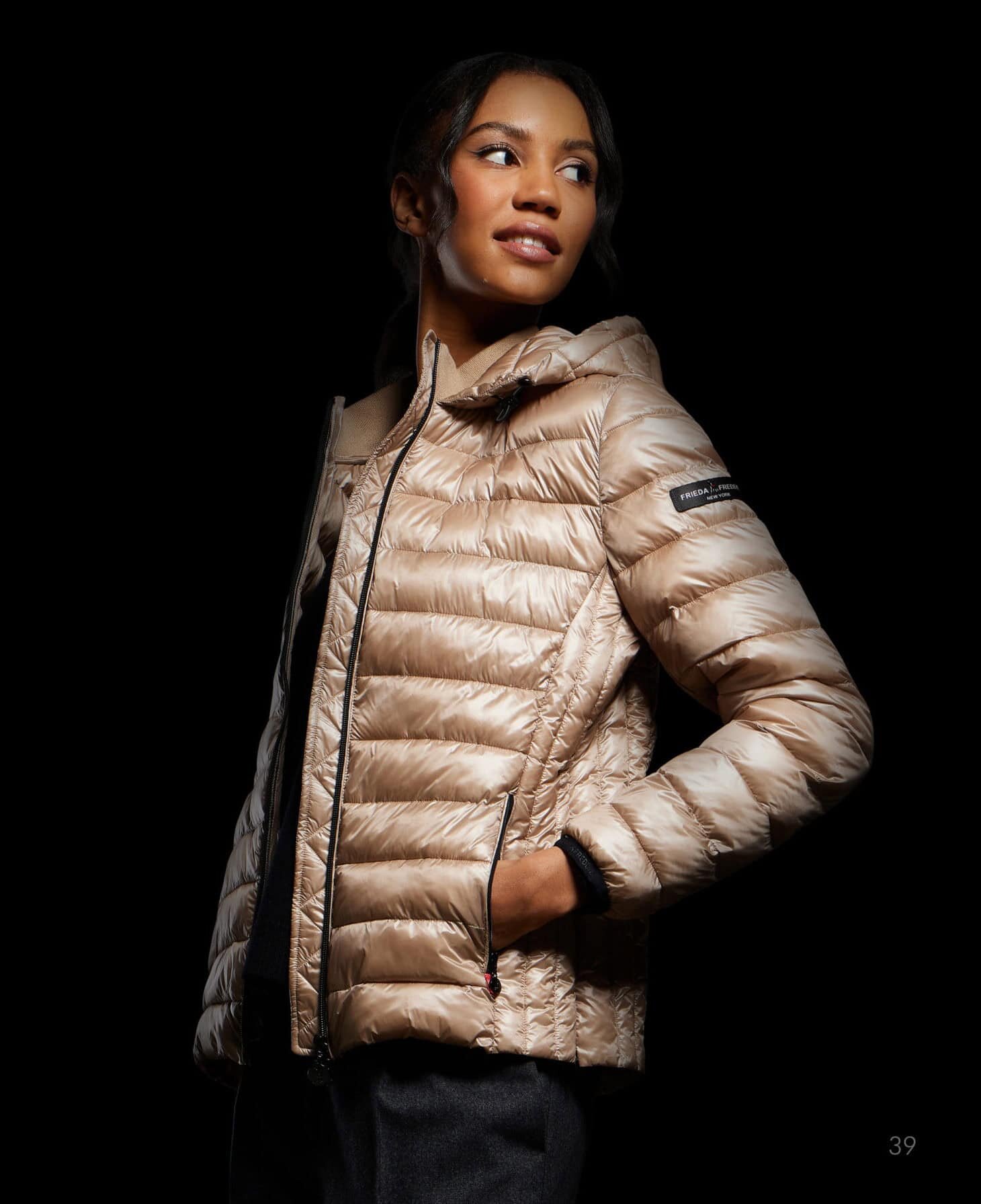 eine frau steht vor einem schwarzen hintergrund, trägt eine glänzende beigefarbene pufferjacke, hat die hände in den taschen, lächelt und schaut zur seite.