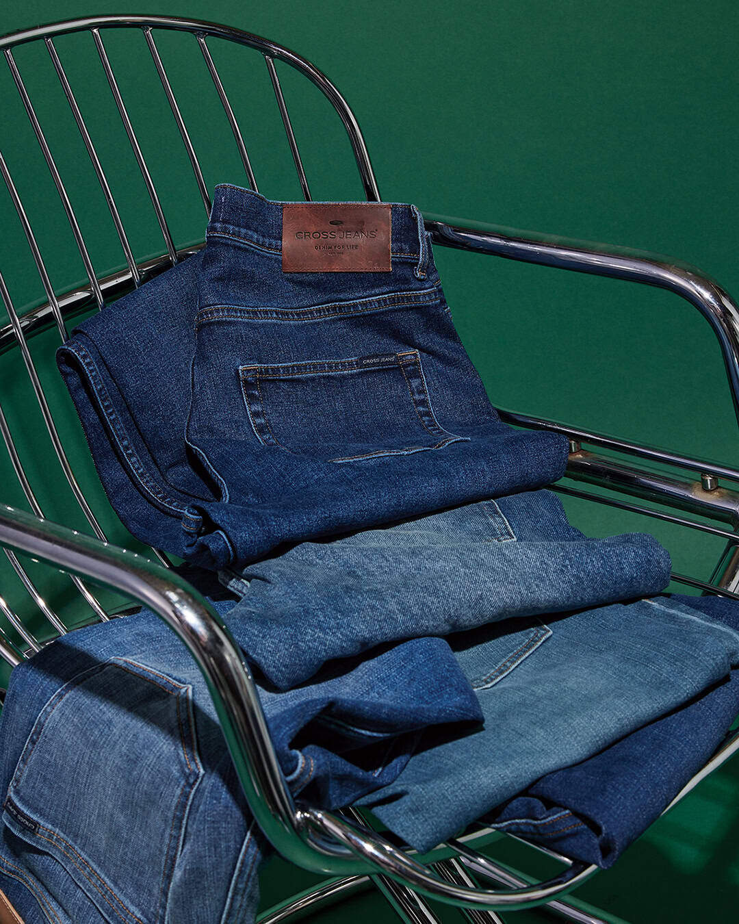 ein stapel gefalteter blue jeans in verschiedenen waschungen sitzt auf einem modernen metallstuhl mit grünem hintergrund. das oberste paar hat einen braunen lederaufnäher am bund.